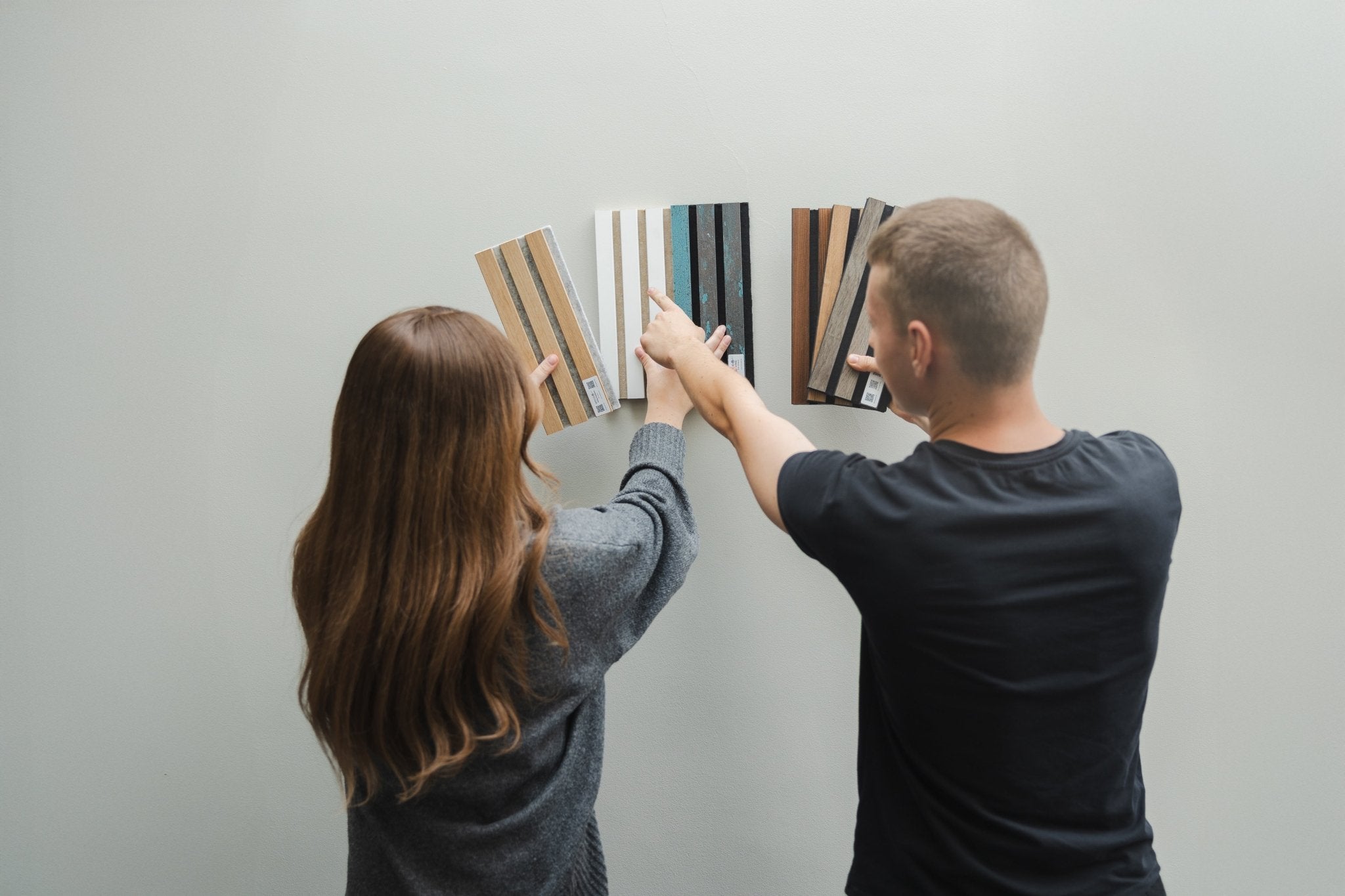 Two people review color and material samples against a light Akupanel acoustic wood panel, considering options for interior design or renovation.